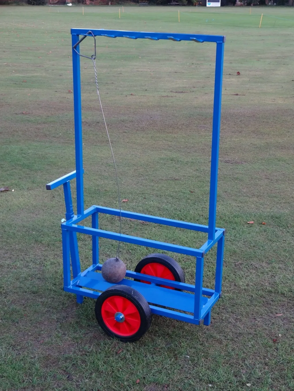 Blue powder-coated steel hammer trolley on grass with two large hard-rubber wheels and a suspended metal hammer under a tall rectangular frame.