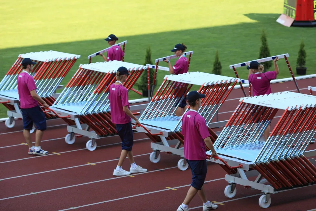 Several silver-anodised hurdle carts holding stacked red-and-silver hurdles being pushed by attendants in pink shirts along a red track.