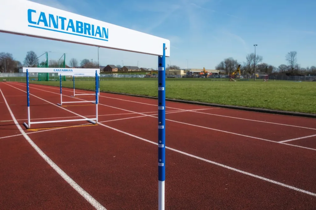 White-topped blue-and-white training hurdles lined on a red running track with a grassy field and equipment in the background.