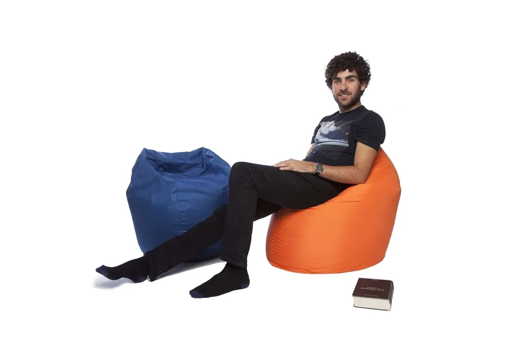 Man sitting on an orange polyester bean bag with a blue polyester bean bag beside him and a closed book on the floor.