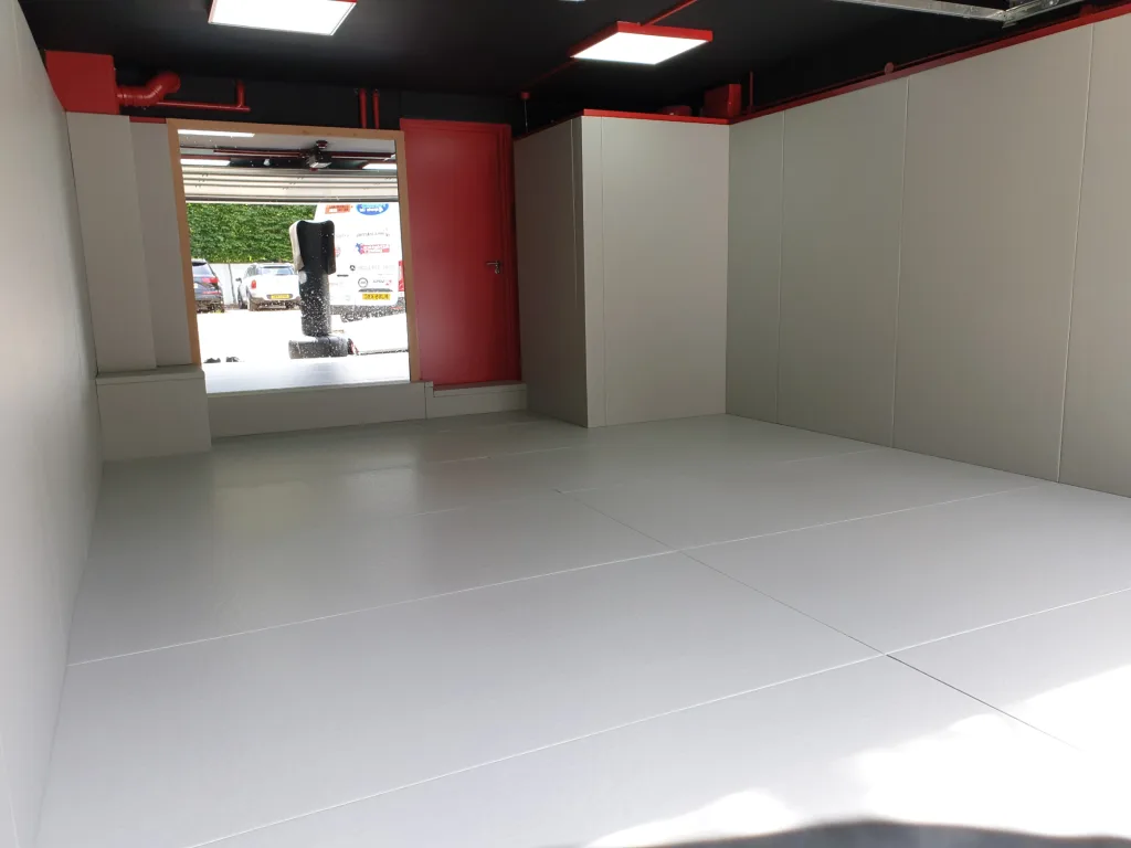 White MMA mats cover the floor of a small training room with light-gray padded walls and a red door at the far end.