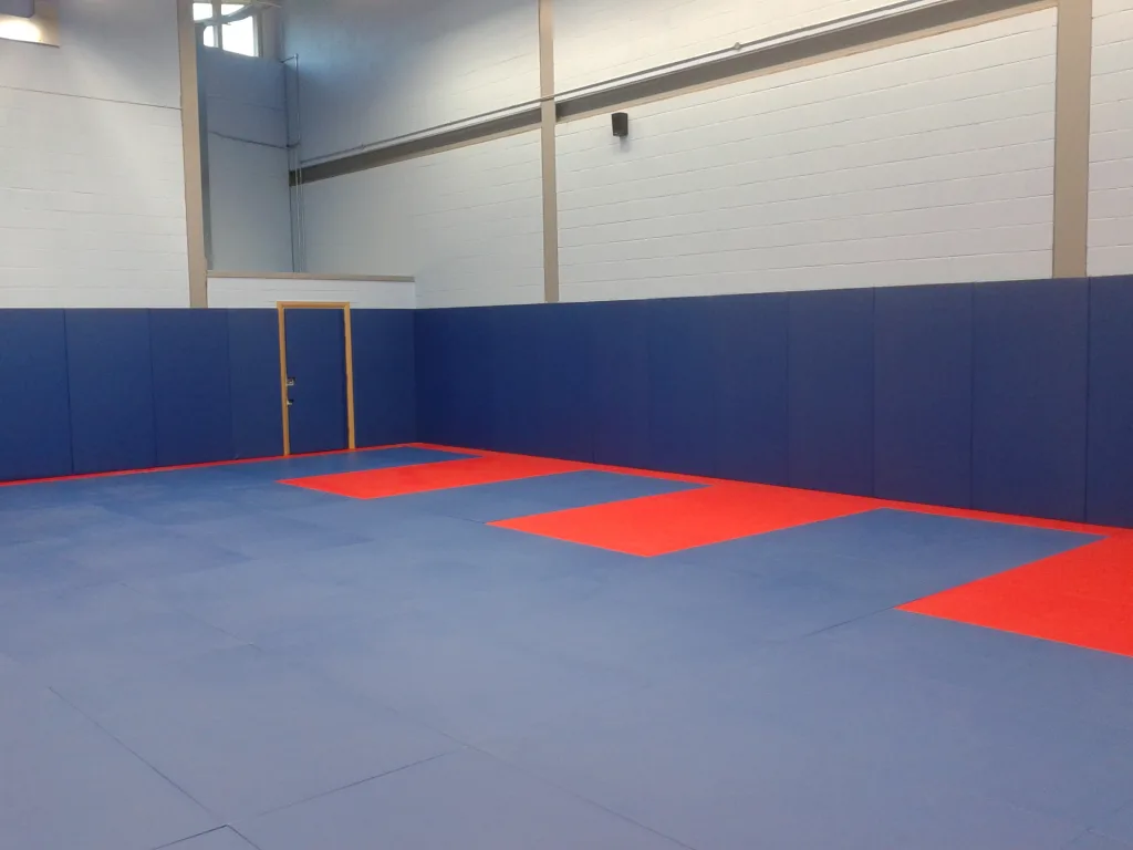 Royal blue and red combat mats cover a gym floor with royal blue wall padding and a yellow-framed door.