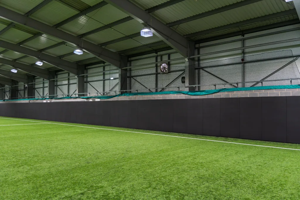 Black sports pitch safety padding made from heavy duty PVC covers mounted along an indoor turf pitch wall beneath metal roof beams and lights.