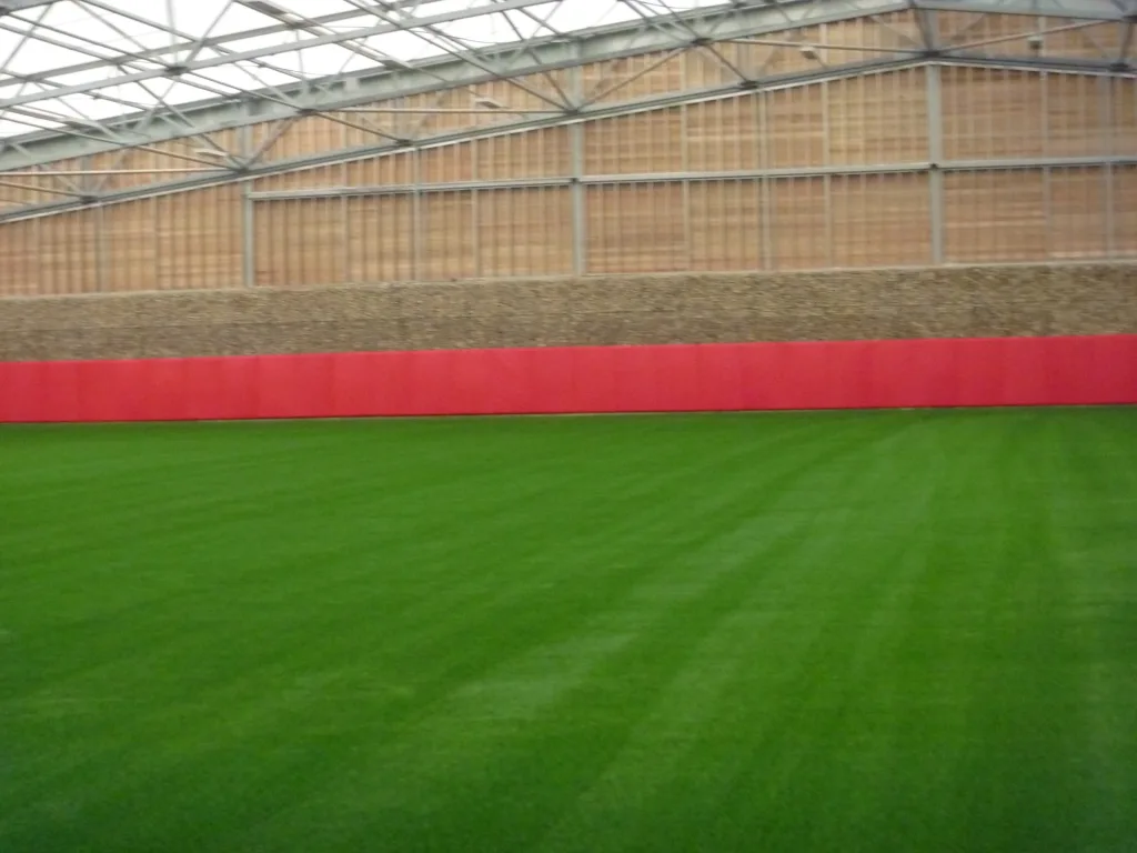 Indoor sports pitch with green artificial turf and red heavy-duty PVC safety padding lining a low wall beneath metal roof trusses.