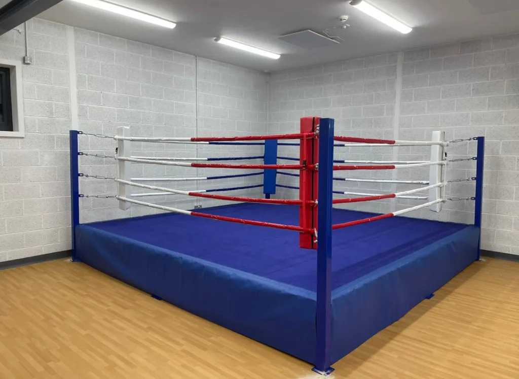 Raised platform training ring with powder-coated steel frame, blue canvas and skirt, red corner pads and red and white ropes in a small training room.