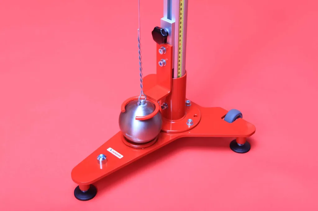 Red metal measuring stand with vertical ruler holding a suspended silver hammer ball by twisted wire over a three-leg base with leveling feet and one caster.