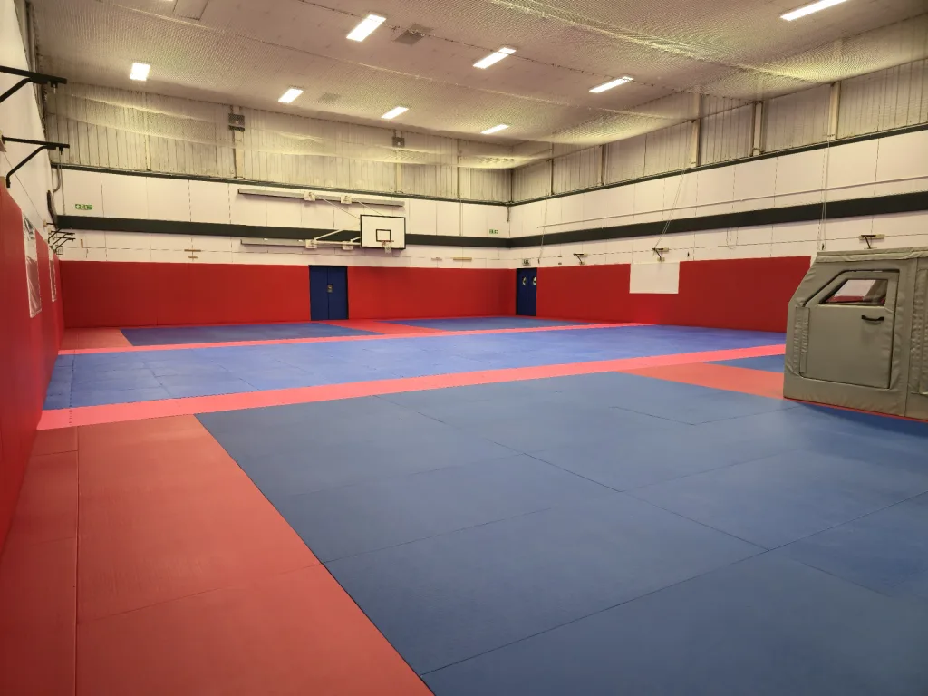 Indoor sports hall with blue tatami PVC judo mats edged by red strips, red wall padding and a mounted basketball hoop.
