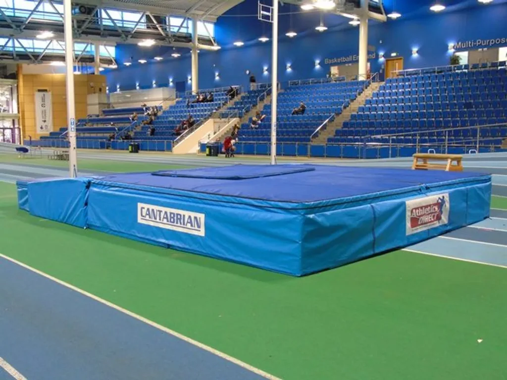 Blue spike-proof push-on mat with foam core placed on an indoor athletics track in front of empty blue spectator seating.
