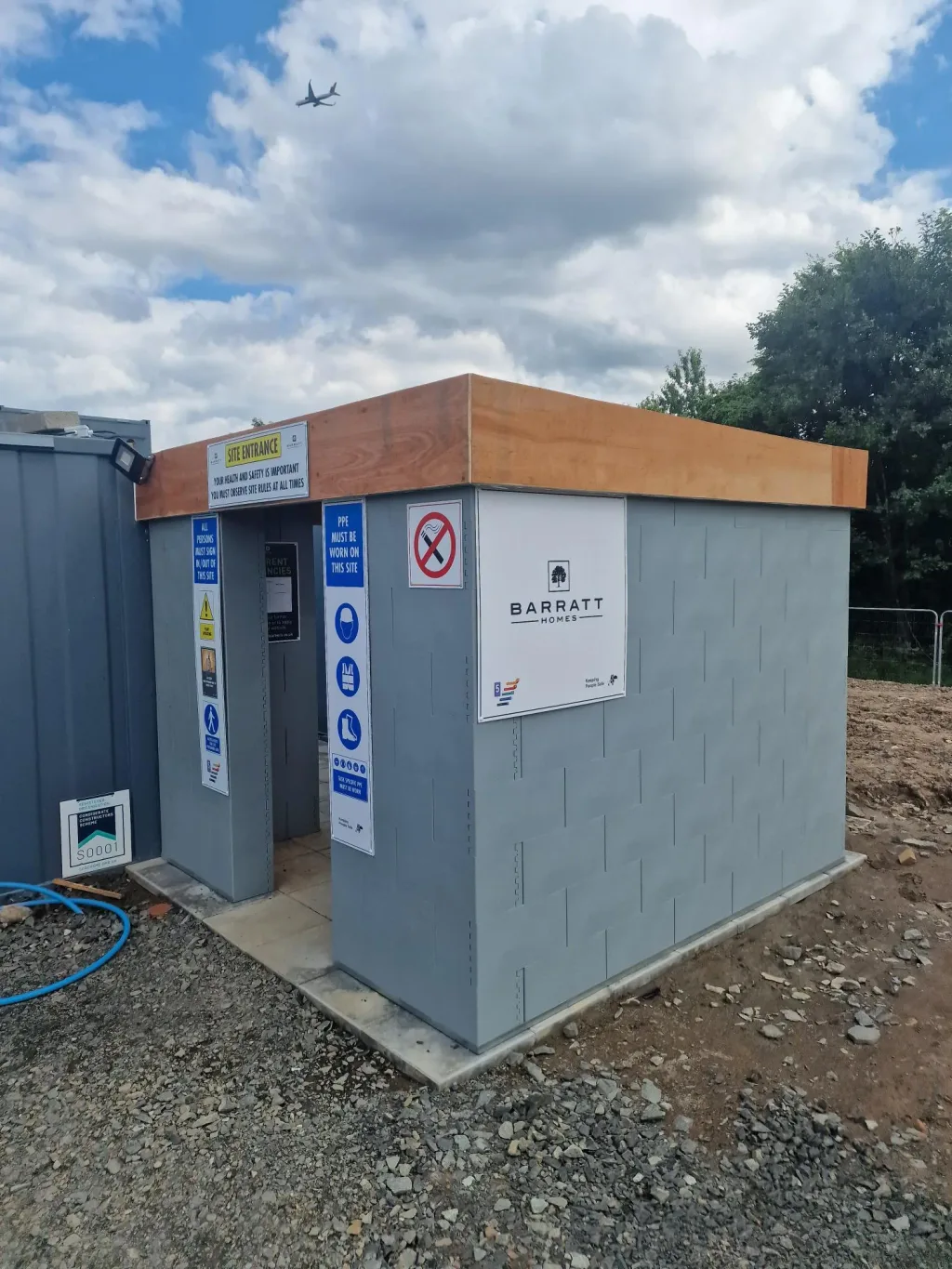 Gray modular training unit with wooden top and safety signage at a gravel construction site under a cloudy sky.