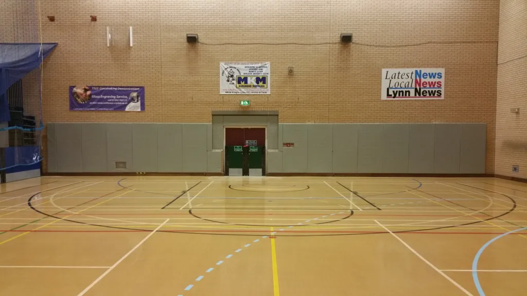 Gym wall lined with grey padded panels, centered double doors, and a sign reading Latest News Local News Lynn News above.