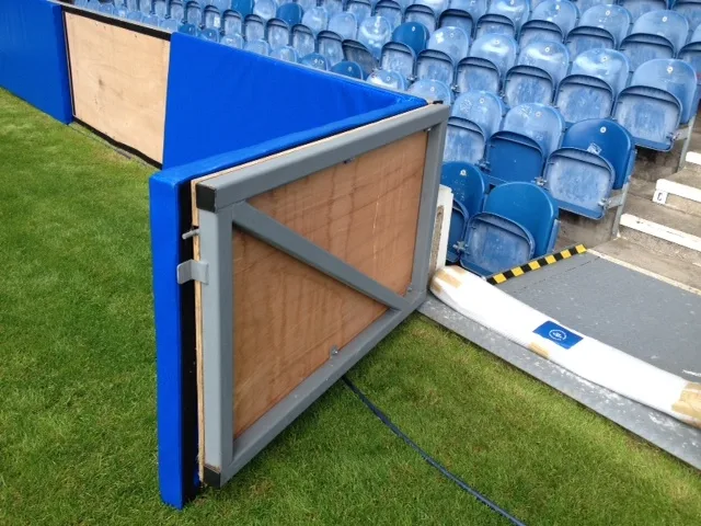 Blue heavy-duty PVC sports pitch safety pad on a metal-framed wooden panel beside grass and blue stadium seats.