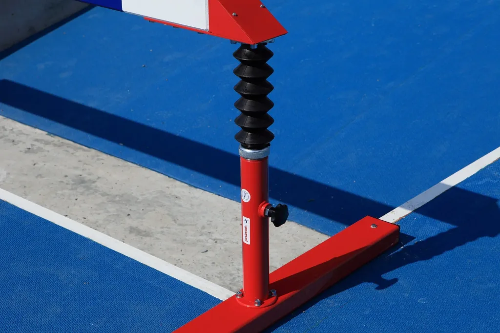 Red telescopic steeplechase hurdle leg with black rubber bellows and Polanik sticker standing on a blue track beside a white lane line.