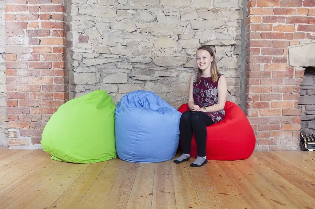Three cotton bean bags in lime, royal blue, and red with a person seated on the red one against an exposed brick and stone wall.