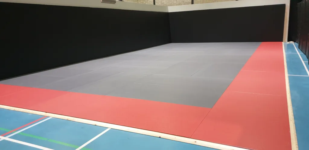 Corner of a training area with interlocking light grey MMA mats bordered by a red edge and black wall pads on a blue sports floor.