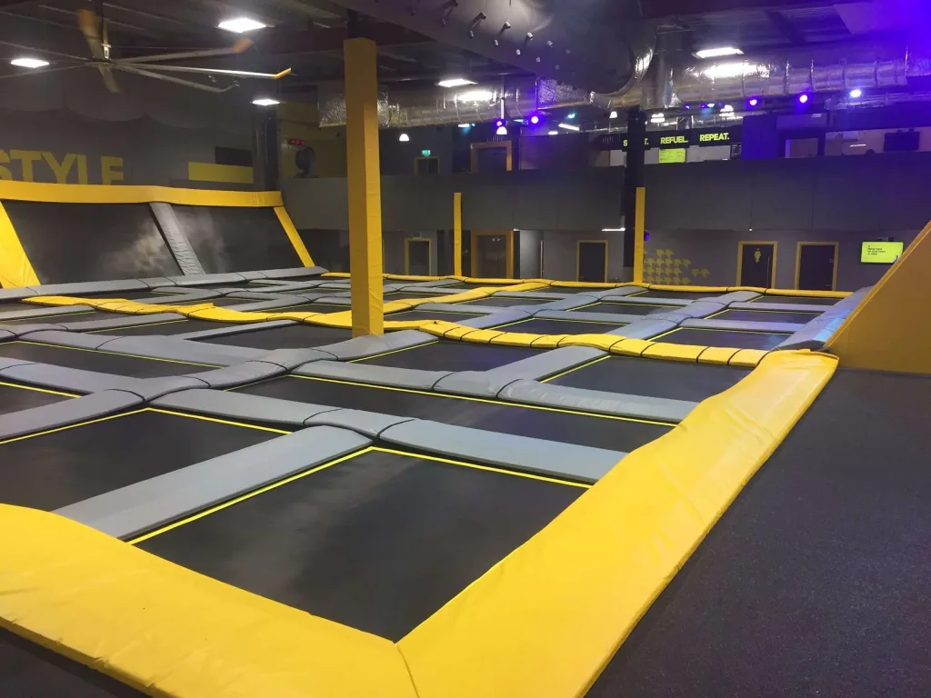 Indoor trampoline park with black jumping mats framed by grey and yellow heavy-duty PVC padding and yellow padded posts.