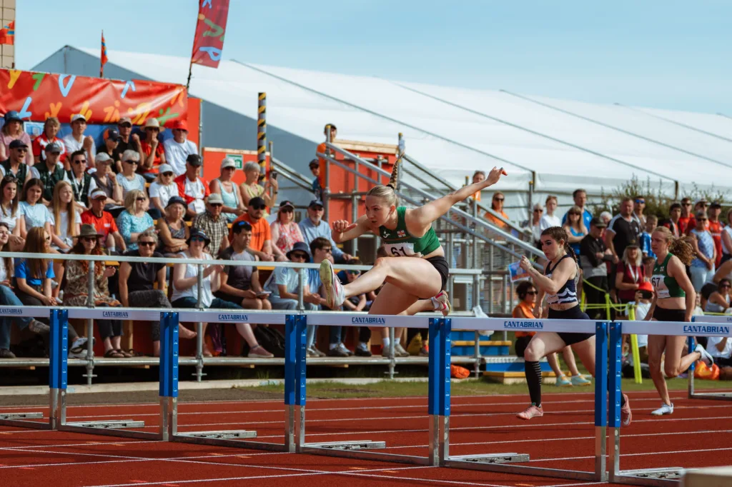 Female hurdler in green midair over a steel hurdle labeled CANTABRIAN with competitors and spectators in the background.
