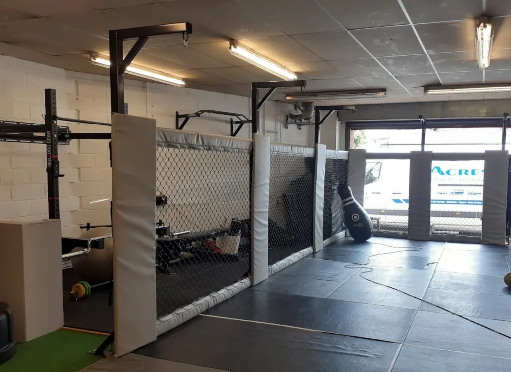 Steel powder-coated cage walling with chain-link mesh and padded panels enclosing gym with freestanding punching bag; van outside reads ACRES.