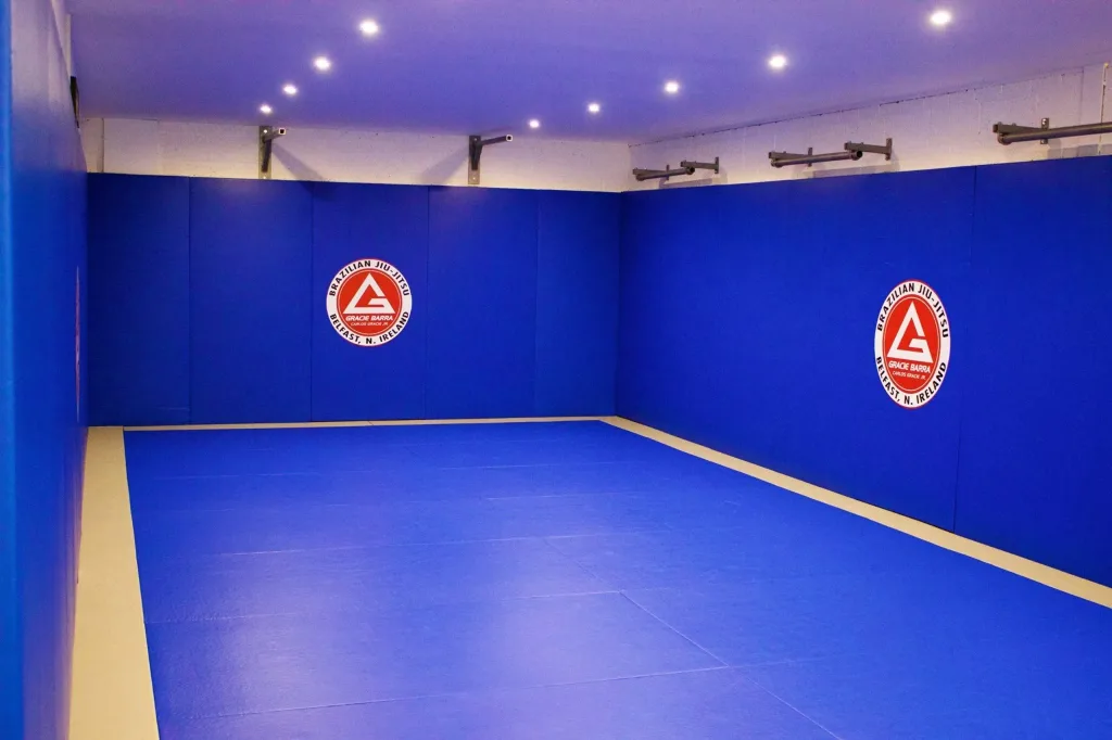 Blue club BJJ mat covering floor and walls with beige border and two red circular logos reading "Brazilian Jiu-Jitsu Gracie Barra Belfast, N. Ireland".