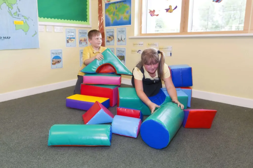 Two children playing with random-coloured soft building shapes on a classroom carpet.