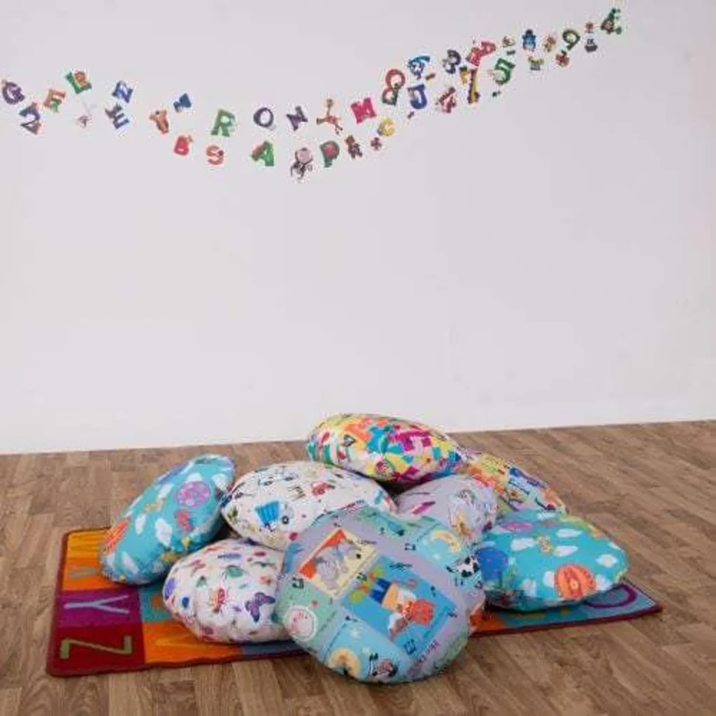 Pile of ten colourful round educational scatter cushions with fibre-filled 100% wipe-clean covers on an alphabet rug against a white wall.