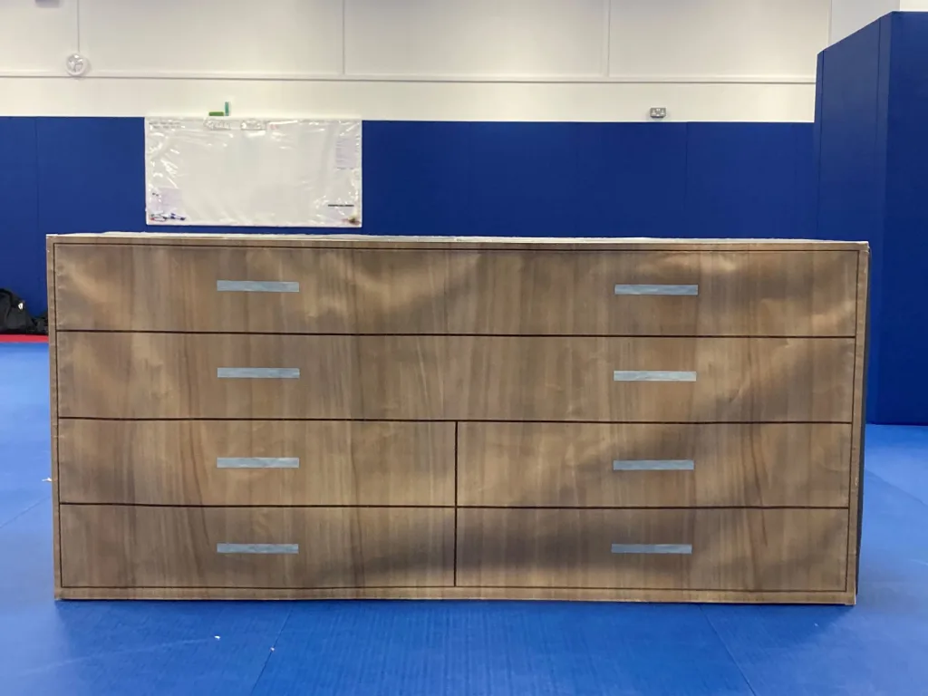 Foam training module printed as a chest of drawers with six faux-wood drawers and silver rectangular handles, on a blue mat in a gym.