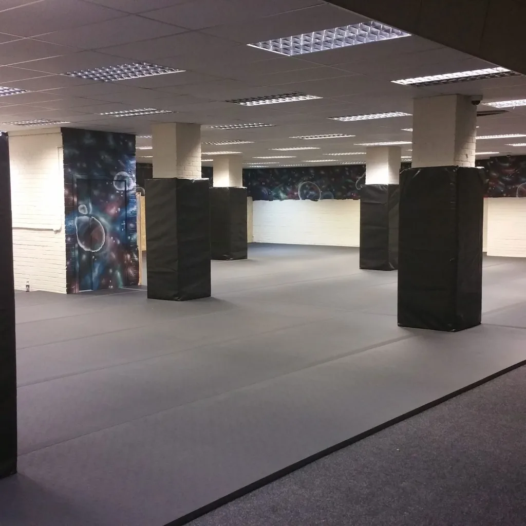 Black square post protectors with heavy-duty PVC covers over foam wrapped around white columns on a gray padded floor in a large indoor room.