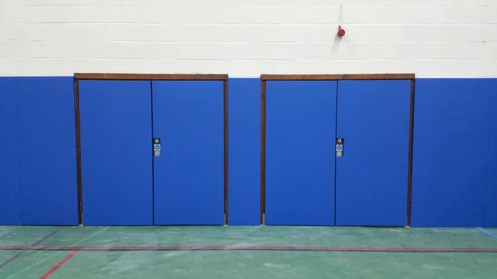Blue wall padding panels covering the lower gym wall with two inset folding doors framed in wood and a red alarm above.