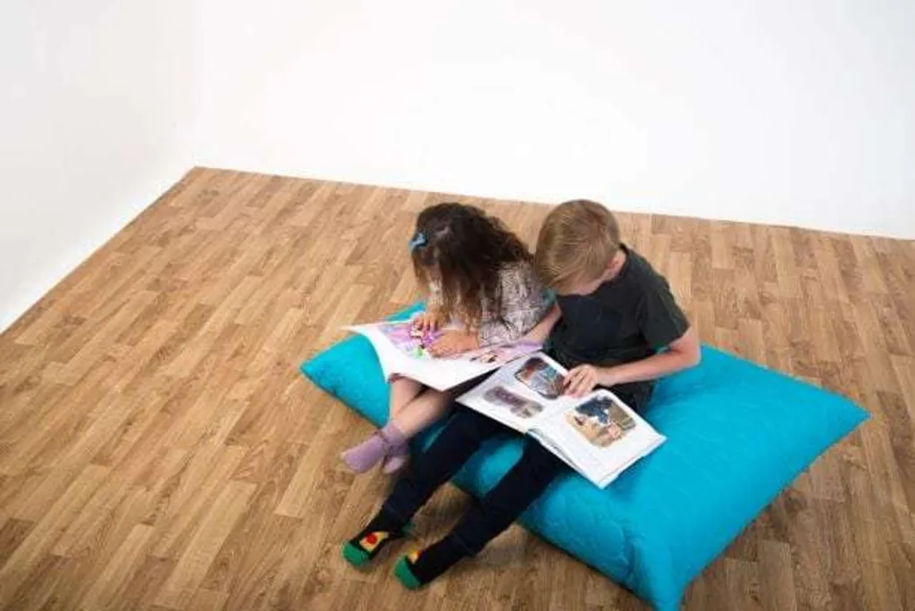 Two children sitting on an aqua padded floor cushion reading books on a light wood floor.