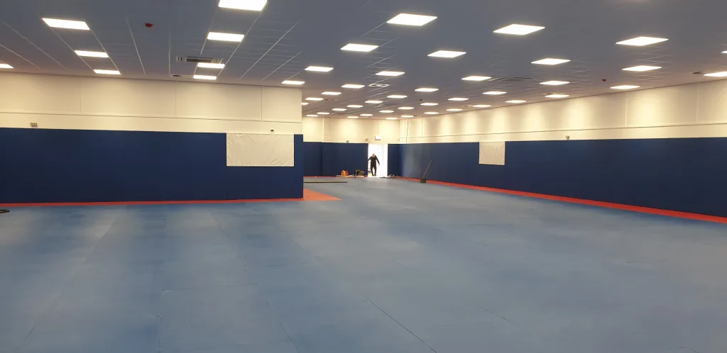 Large indoor dojo with blue tatami-embossed PVC judo mats with red border, blue padded wall panels, overhead lights and a person by the exit.
