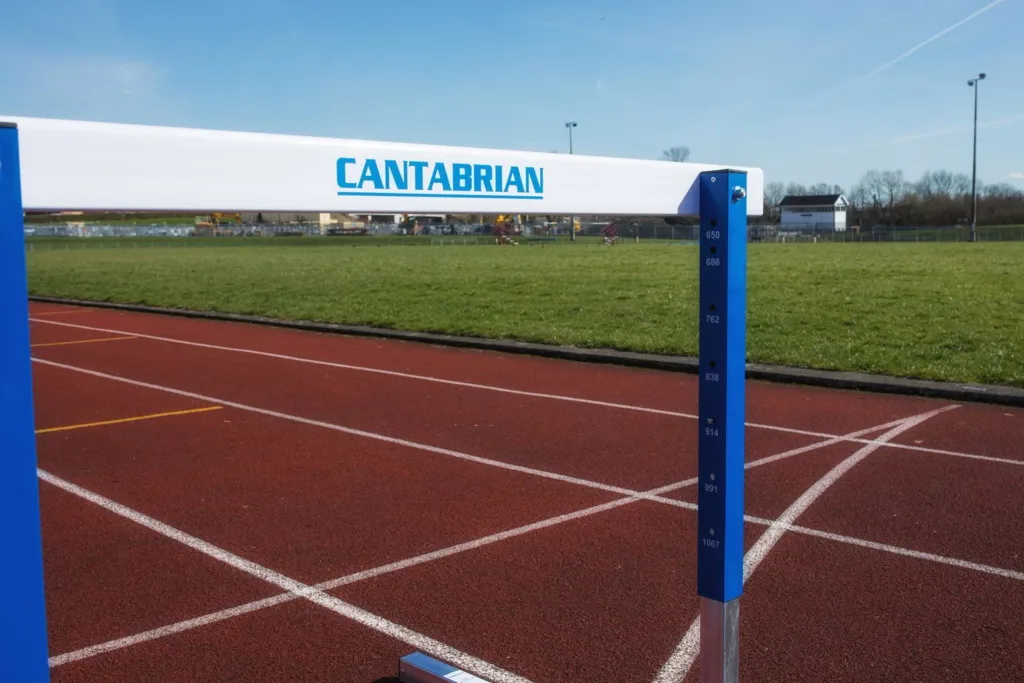 White steel competition hurdle with blue uprights and the text CANTABRIAN on the crossbar, positioned on a red running track.