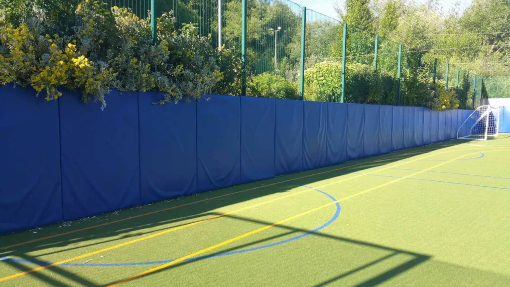 Green artificial sports pitch with a long blue padded side wall topped by shrubs and green mesh fencing, marked by yellow and blue lines and a white goal at right.