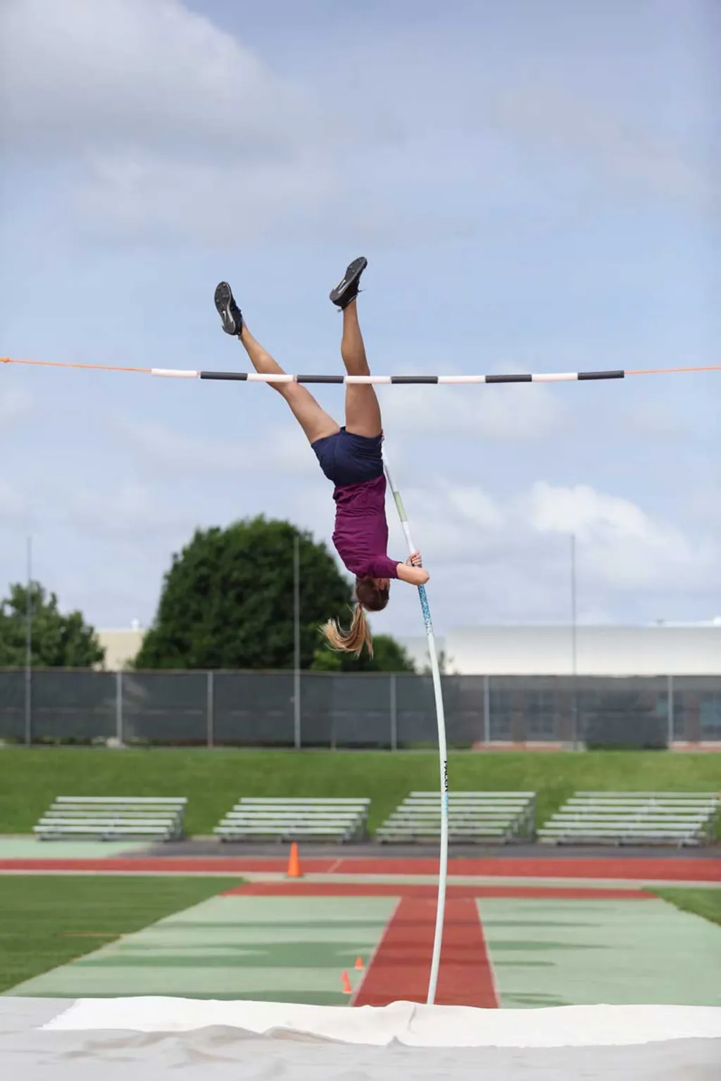 Pole vaulter inverted above the crossbar, gripping a bent pole while hovering over the landing mat on an outdoor track.