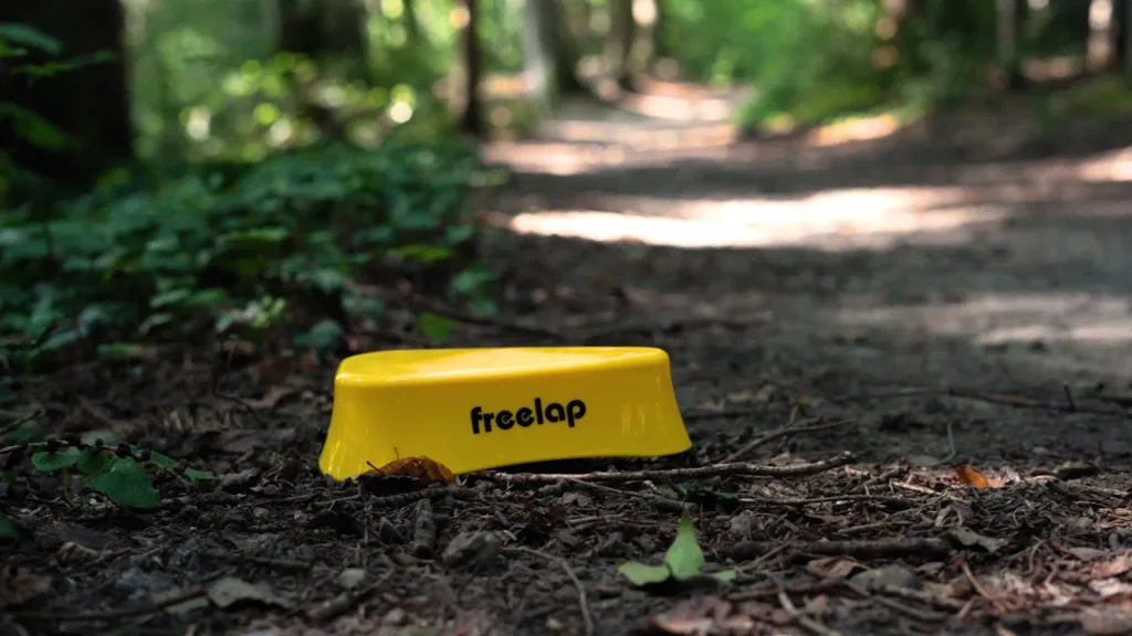 Yellow timing transmitter labeled freelap resting on a leaf-strewn forest trail.