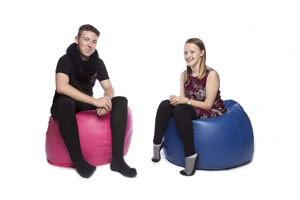 Two young adults seated on teen/adult faux leather bean bags in hot pink and royal blue, smiling on a white background.