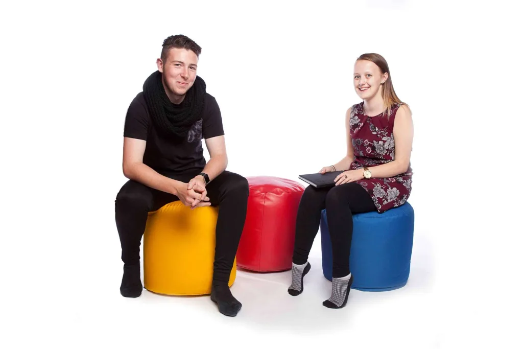 Two people seated on faux leather bean pouffes, a man on yellow and a woman on blue with a red pouffe between them.