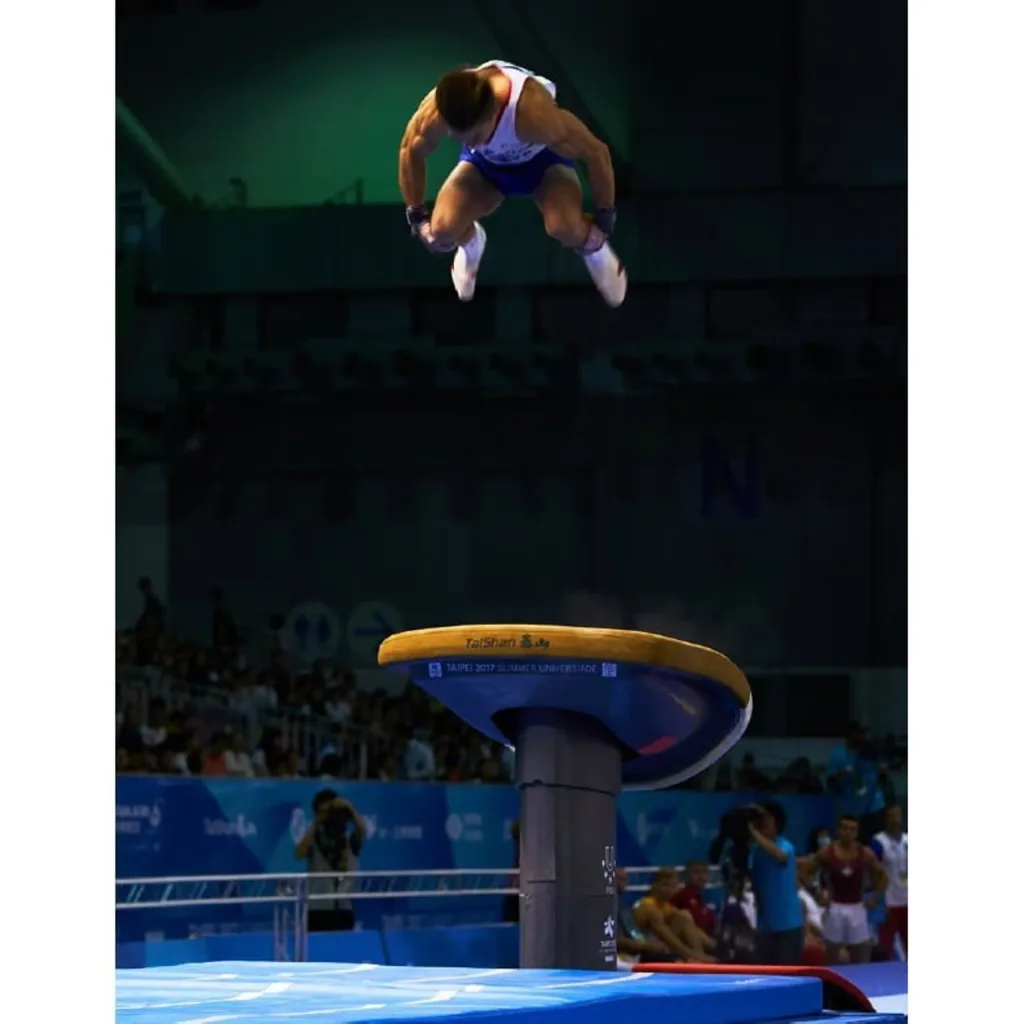 Male gymnast tucked midair above a vaulting table labeled Taishan, with a blue landing mat and spectators in the arena.