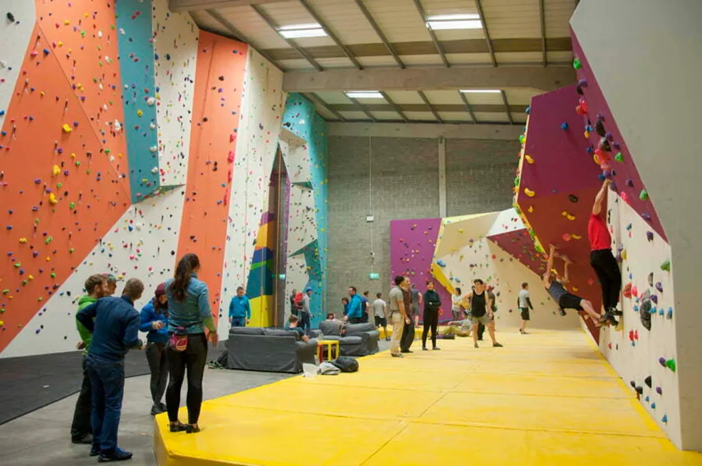 Indoor climbing gym with yellow double-sided PVC bouldering mats covering the floor and climbers on colorful angled walls.