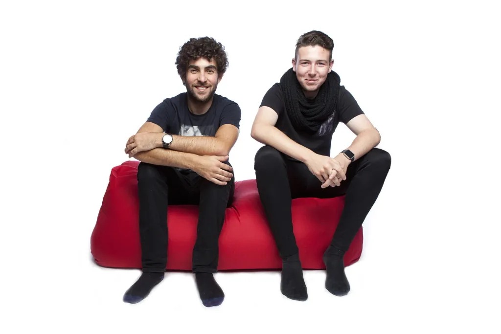 Two young men seated side by side on a red polyester bean bench, smiling toward the camera.
