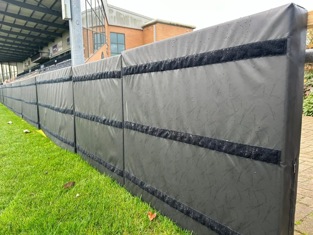 Padded barrier panels along a sports pitch covered in sewn UV- and tear-resistant heavy-duty PVC with horizontal hook-and-loop strips and raindrops.