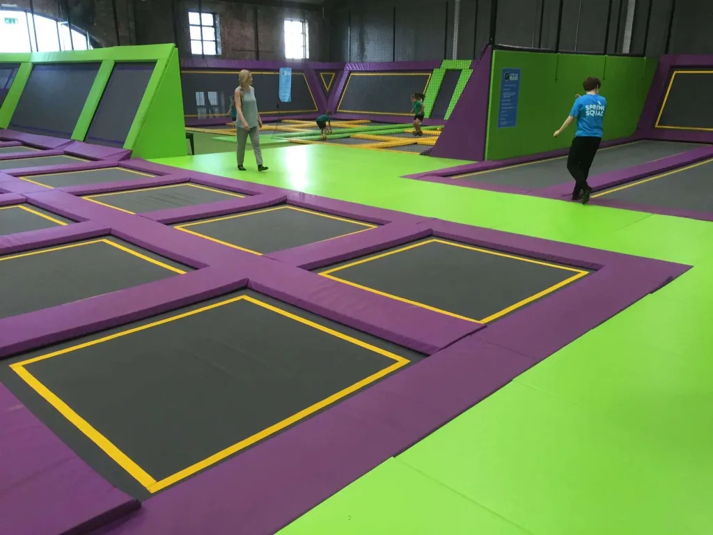 Green and purple PVC-covered trampoline park padding with black jumping mats outlined in yellow, and a person in a blue shirt reading SPRING SQUAD.