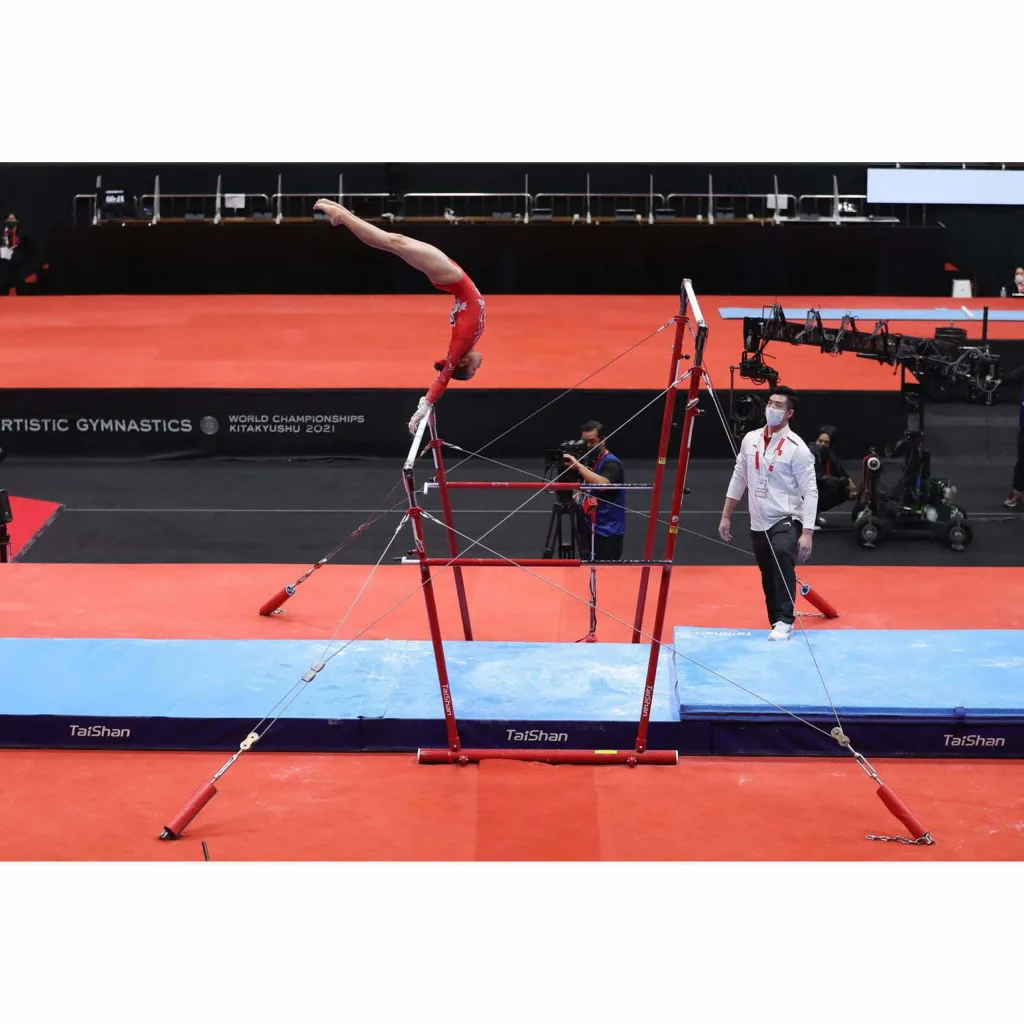 Gymnast in a red leotard performing a handstand on red uneven bars above blue landing mats, with "WORLD CHAMPIONSHIPS KITAKYUSHU 2021" visible.