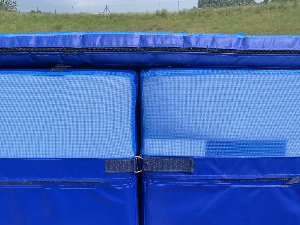 Blue PVC-covered pole vault landing mats showing two interlocked modules with zippers, carry handles and a central metal D-ring, grassy field beyond.