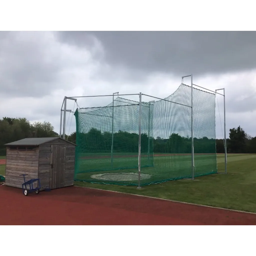 Discus cage with green knotless net on galvanised steel poles around a throwing circle next to a wooden shed on a track under cloudy sky.