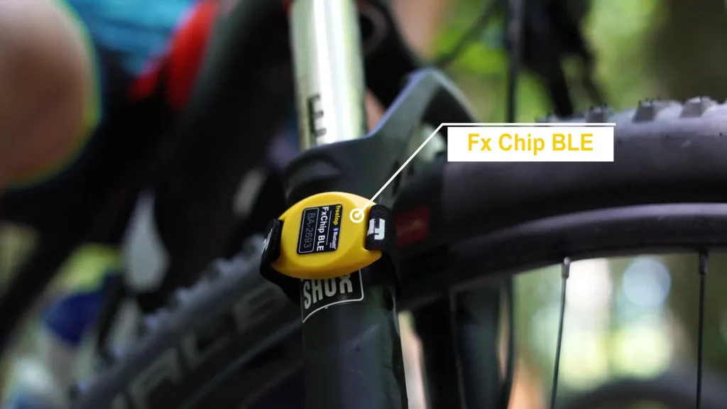 Yellow FxChip BLE with a black clip attached to a bicycle fork near the wheel, label reads 'FxChip BLE BA-2693' and overlay reads 'Fx Chip BLE'.