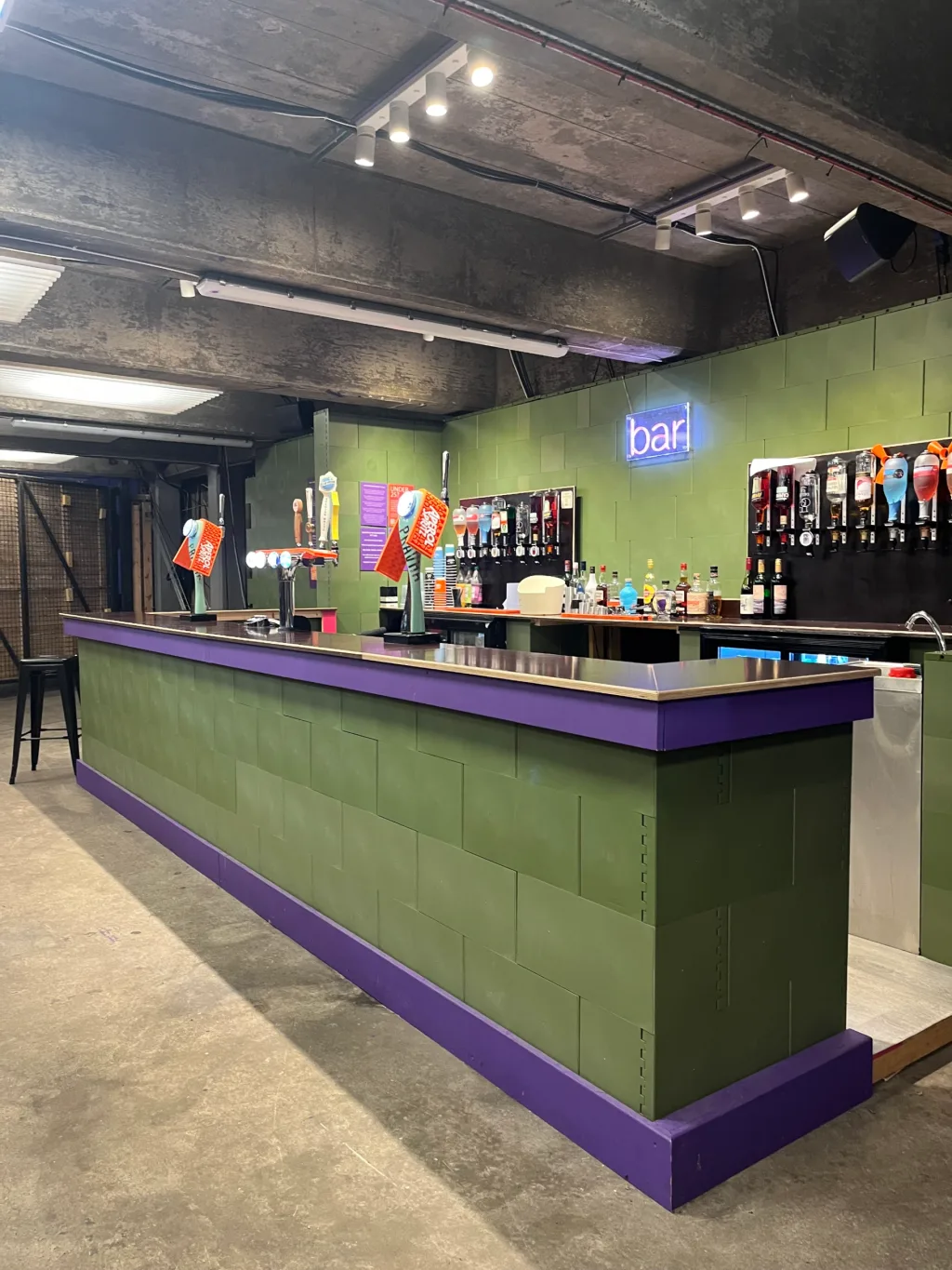 Long green modular bar with purple trim and stainless countertop, beer taps and bottles behind it and a neon sign reading bar.