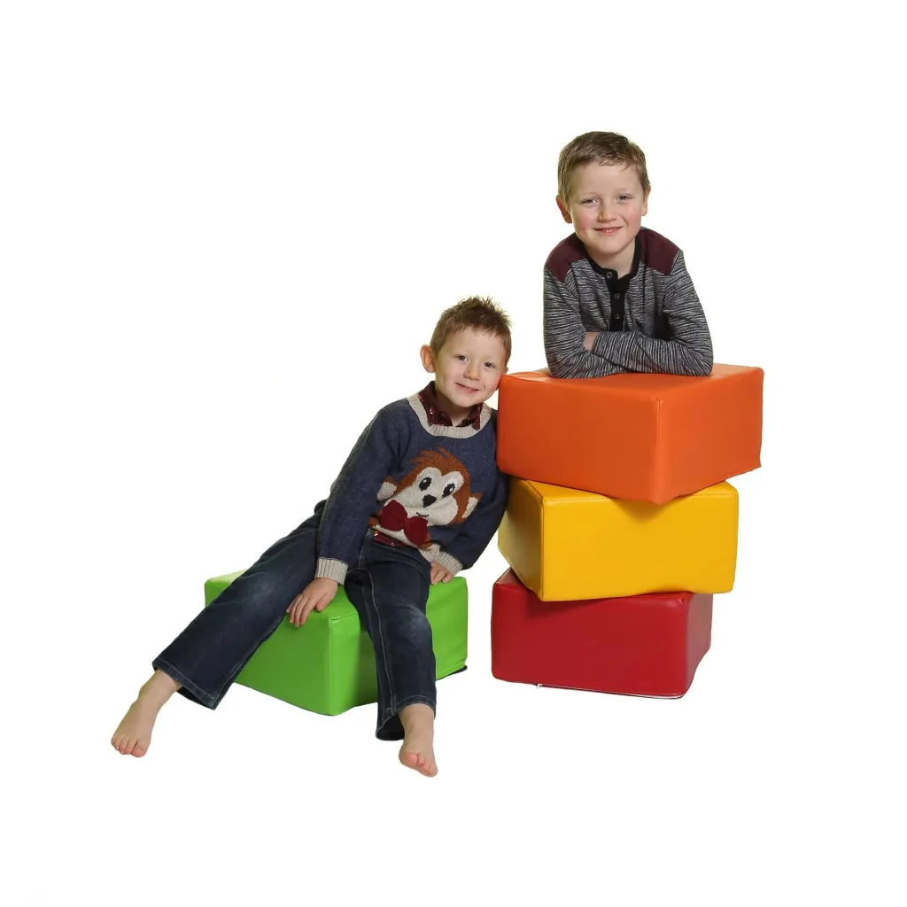 Two smiling boys sit on and beside a stack of five cotton-covered square foam seating pods in green, red, yellow and orange.
