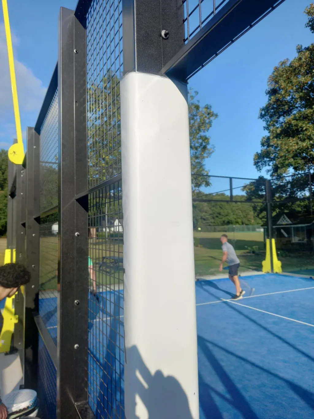 Padel court metal post wrapped in PVC-covered foam padding, mesh wall in front and players on the court in the background.