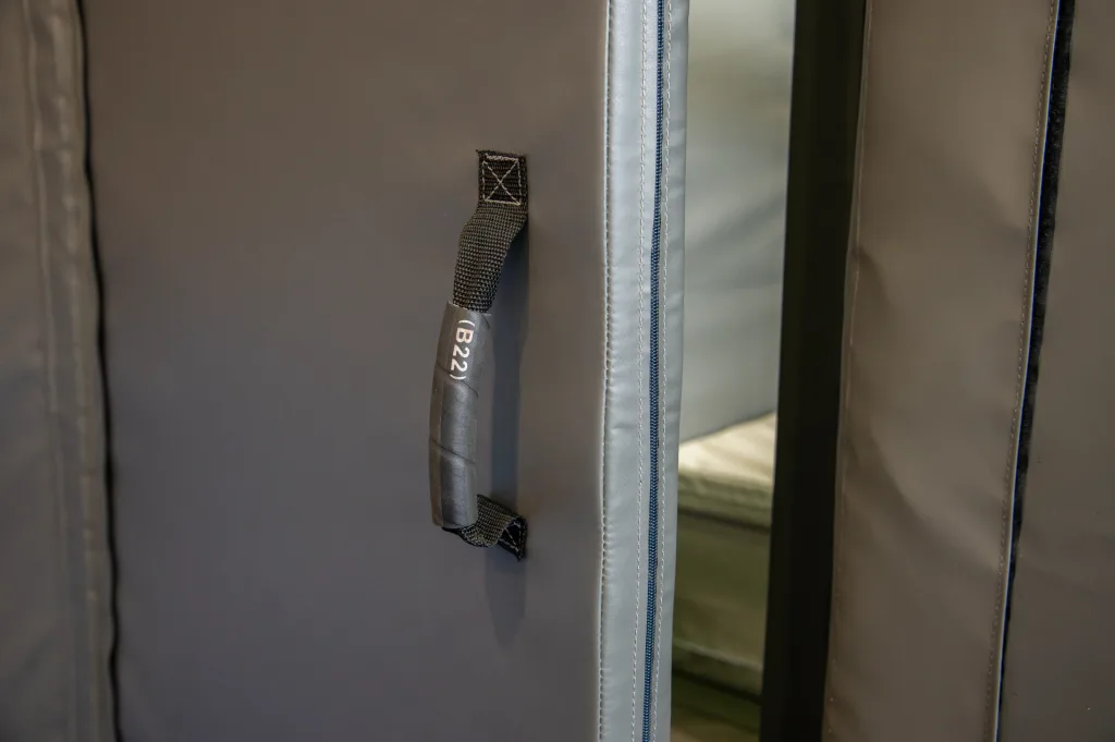 Close-up of a grey padded PVC custody cell wall with a black webbing handle labeled (B22) and a light-gray zip seam.