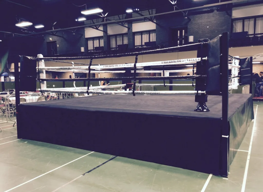 Platform training ring with a powder-coated steel frame, raised deck, black canvas and skirts, and white ropes in an indoor sports hall.
