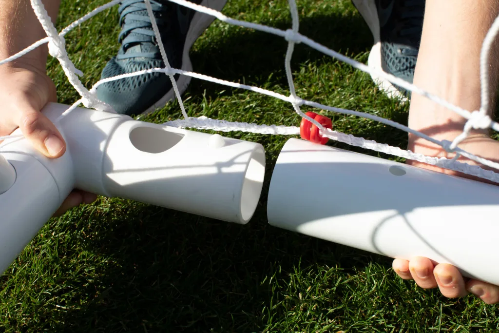 Hands connecting two white plastic goal frame tubes beneath a white net with a red clip on green grass.